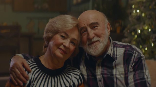 Portrait Of Loving Senior Couple Sitting Close Together At Home, Cuddling Caringly, Smiling And Looking Into Camera. Decorated Christmas Interior In The Background. Shot On ARRI Alexa Mini With Cooke 