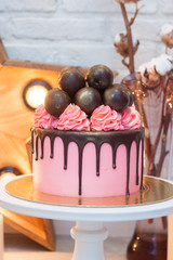 Pink cake decorated with chocolate cake pops, cream cheese swirls and chocolate sauce on white cake stand. Wooden star, burning candles and cotton flowers on the background. 
