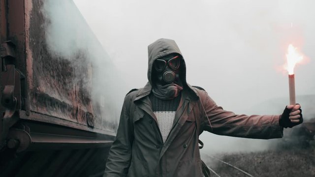 Hooded man in protective gas mask walks through abandoned carriages at destroyed train station holding burning red signal fire in hand. Apocalypse, survivor after nuclear or chemical disaster