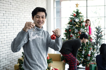 Young Asian man holds the toy to decorate the Christmas tree with smile on which has friends...