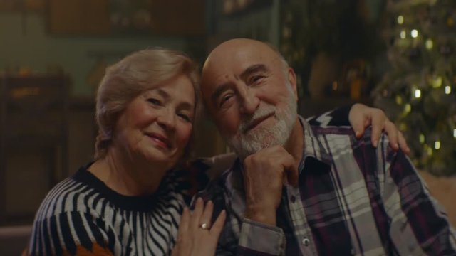 Portrait Of Loving Senior Couple Sitting Close Together At Home, Cuddling Caringly, Smiling And Looking Into Camera. Decorated Christmas Interior In The Background. Shot On ARRI Alexa Mini With Cooke 