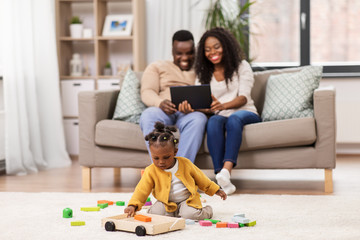 childhood and people concept - little african american baby girl playing with toy blocks and parents with tablet pc computer at home