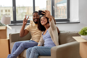 Obraz premium moving, repair and real estate concept - happy african american couple with cardboard boxes sitting on sofa and taking selfie by smartphone at new home