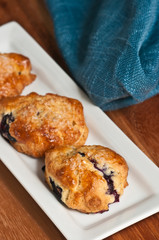 Top view, close distance of three freshly baked, warm blueberry scones of a white, rectangular plate with a blue, cloth napkin on a wood serving board