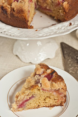 Top, front view, close distance of a wedge of plum, sour cream, coffee cake on a round, white plate  and a pedestaled, display dish with res of the coffee cake and a silver pie server