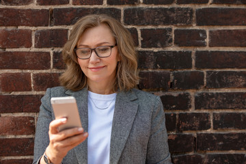 Portrait of a happy beautiful blonde woman, 55 years old, smiling, with a smartphone in hand. Outdoors, in the city.
