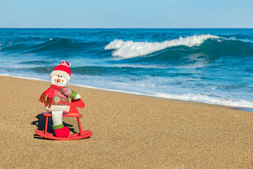 Snowman figurine on the beach, tropical ocean background, sand and waves, Christmas travel concept, copy space