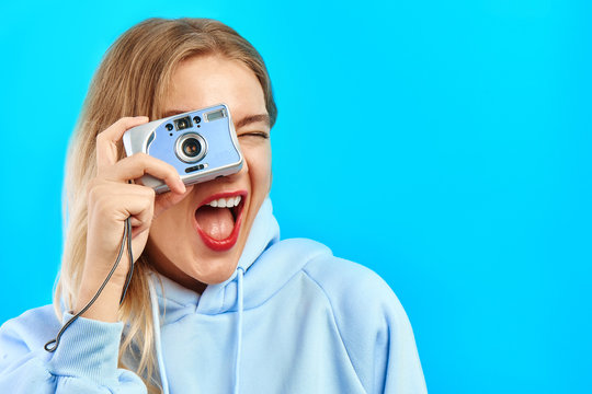 Portrait of young positive hipster photographer girl taking pictures on film camera, smiling cheerfully on blue background with copy space
