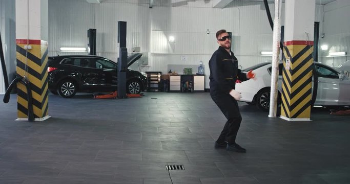 Charismatic smiling large mechanic man in uniform and protected glasses dancing funny after his finished the work day in a modern service auto center