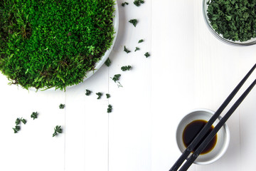 A plastic plate with green moss stands on a white wooden table and next to it are containers with dried parsley and soy sauce. And next to it are black chopsticks.