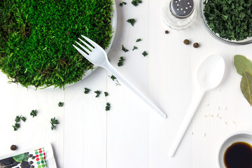 A plastic plate with green moss stands on a white wooden table and next to it are containers with dried parsley soy sauce and a salt shaker. And on the side are a disposable fork with a spoon.