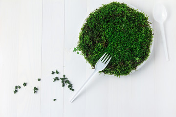 A round plastic plate with green moss stands on a white wooden table and next to it is scattered dried parsley and a disposable fork.