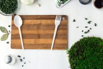 A round plastic plate with green moss stands on a white wooden table and next to it is scattered dried parsley and a disposable spoon with a fork on a cutting board.