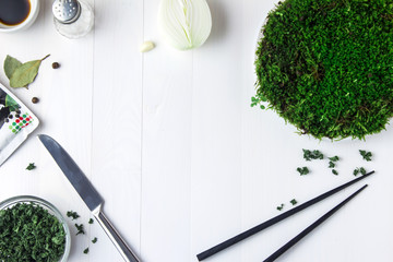 A round plastic plate with green moss stands on a white wooden table and next to it is scattered dried parsley and a knife with black chopsticks lies.