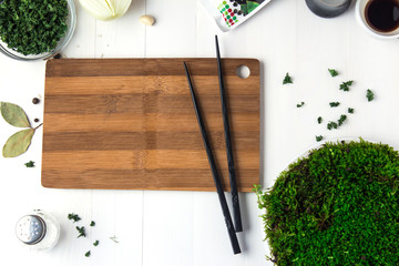 A round plastic plate with green moss stands on a white wooden table and next to it is scattered dried parsley and black chopsticks lie on a cutting board.