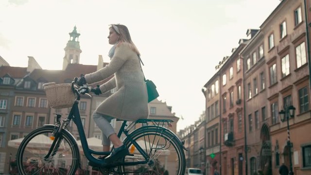 Woman in Coat Rides Bicycle in City Square with Old Buildings in Cold Autumn Day. Pretty Lady on Retro Bike. Urban Cycle Chic and European Architecture. 4K Medium Tracking Orbit Shot, Backside View
