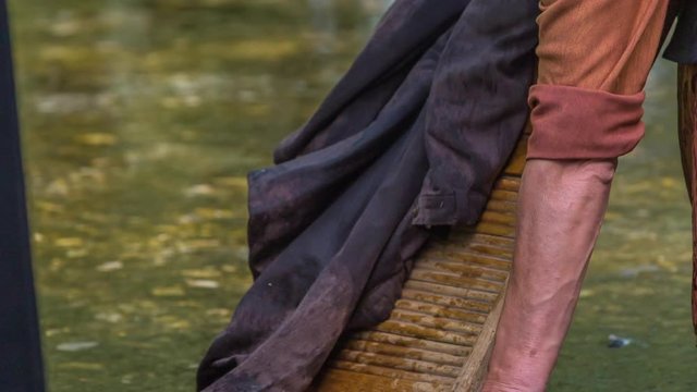 An older woman is wahsing clothes on a wooden board in a river and she is using a brush to get rid of the stains.