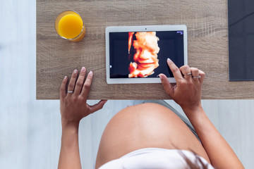 Pregnant woman looking and touching the digital tablet with the ultrasound of her baby at home.