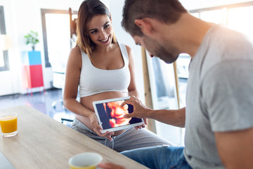Pretty young pregnant holding the digital tablet with the ultrasound of her baby while husband...