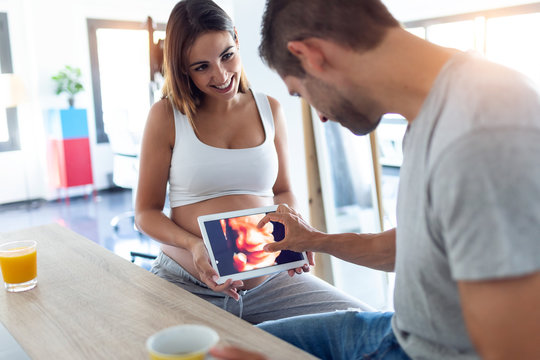 Pretty young pregnant holding the digital tablet with the ultrasound of her baby while husband looking this at home.