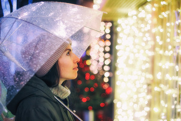 Portrait of young beautiful girl holding umbrella with garland. Posing in Christmas fair, in street of european city. Snowfall. Model wearing knitted sweater, scarf, hat. Christmas mood