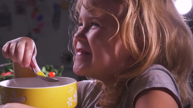 Cute little girl eating cornflakes from a yellow cup in the kitchen. Happy funny smiling baby face. Healthy babies food. Happy child eating. Children nutrition. Lunch or dinner, breakfast at home.