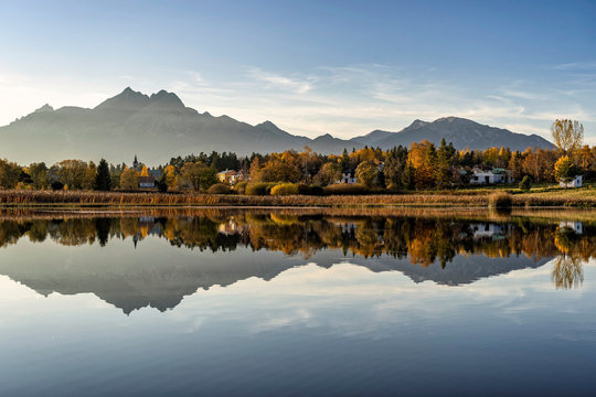 Peaceful scene of beautiful autumn mountain landscape with small settlement, calm lake, colorful trees and high peaks in High Tatras, Slovakia.
