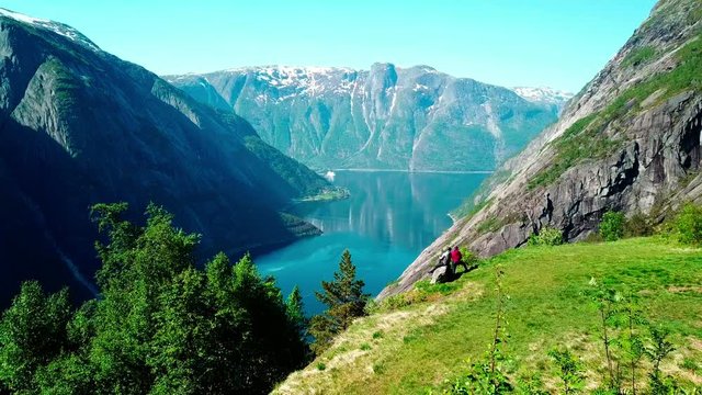 A drone areal capture of a fjord in Norway. Flight over a couple standing at the edge of the mountain. Long view on the fjord. Beautiful clarity in the sky. Lush green slopes, deep blue color of water