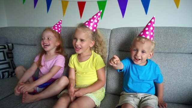 Laughing kids with party hats watch tv. Little boy and two girls watching comedy