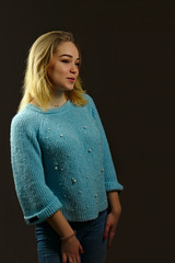 A blonde girl in jeans and a blue sweater stands on a dark background and smiles shyly
