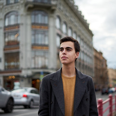 Obraz premium Handsome stylish fashionable man, brunette in elegant gray coat, stands on street in historical center of St. Petersburg. Young man with dark hair, thick eyebrows.
