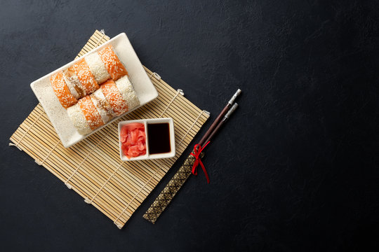 Sushi roll, chopsticks, soy sauce, ginger and bamboo mat on black background. Top view with copy space. Flat lay.