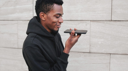 Portrait modern african man holding in hands smartphone using voice command recorder, assistant or takes calling, looking at phone on city street on gray wall background