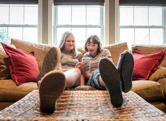 Two teenage girls sitting on couch with feet up looking at cellphone.