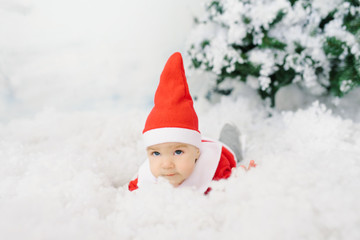 A five-month-old baby in a little Santa costume lies on artificial snow under a Christmas tree.