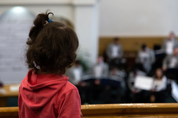 A view from the back of a little girl in a red sweater, who is looking at an orchestra that is out of focus. © lunarts_studio