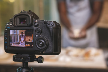 Baking process. Young cooking african man speaker talking on digital camera recording vlog. African male vlogger or shooting blog filming reportage advertising video, coaching.