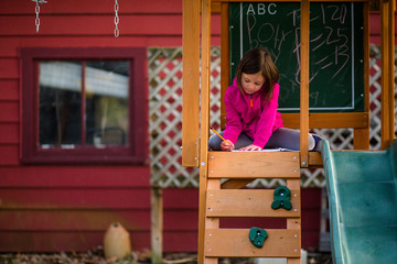A little girl sits in a playset writing in a notebook with a pencil
