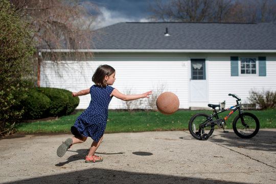 A smal girl chases a basketball in driveway against cloudy sky