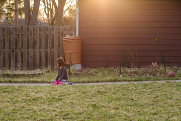 A focused small girl sits on a path in yard at sunset writing a letter
