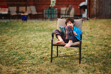 A happy boy reads a book barefoot in backyard in cool weather