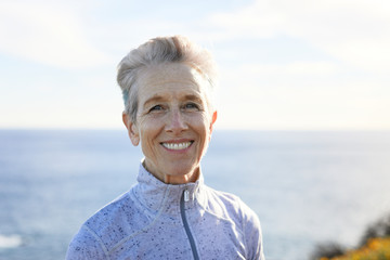 Portrait of smiling senior woman standing at beach against sky during sunny day