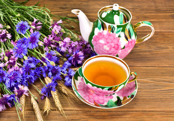 Black tea in a cup with painted teapot, blue cornflowers and wheat ears on a wooden background.