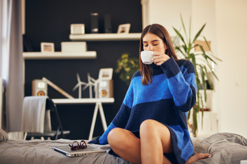 Charming brunette in blue sweater sitting in bedroom in morning and drinking coffee.