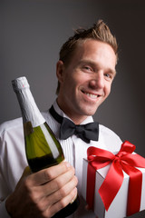 Smiling young man with funky hair and black tie tuxedo shirt holding a bottle of bubbly and gift with romantic red satin ribbon
