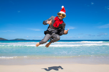 Excited businessman in Santa hat, sunglasses and big red Christmas bow jumping on the shore of a tropical beach