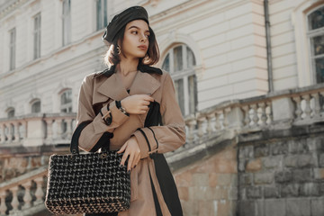 Outdoor autumn fashion portrait of young elegant lady wearing trendy faux leather beret, classic beige trench coat, holding black tweed bag, handbag. Copy, empty space for text - obrazy, fototapety, plakaty