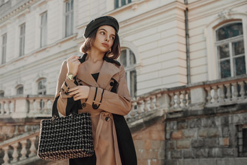 Outdoor autumn fashion portrait of young elegant lady wearing classic beige trench coat, trendy faux leather beret, holding black tweed bag, handbag, posing in street of European city. Copy space 