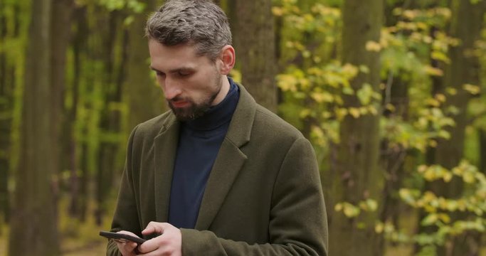 Portrait of a handsome gray-haired Caucasian man in the forest with the smartphone, shaking his head and taking picture of surroundings. Cinema 4k footage ProRes HQ.