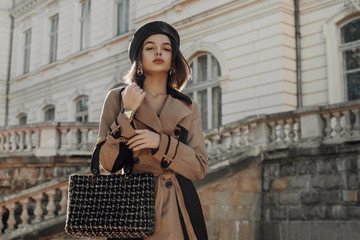 Outdoor autumn fashion portrait of young elegant woman wearing classic beige trench coat, trendy faux leather beret, holding black tweed bag, handbag, posing in street. Copy, empty space for text - obrazy, fototapety, plakaty